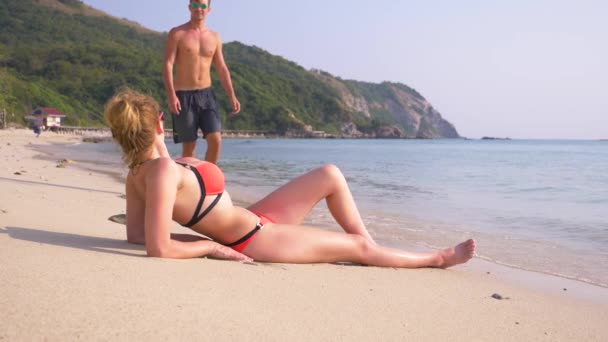 4К, замедленная съемка. Женщина лежит на пляже, к ней подходит красивый молодой человек — стоковое видео