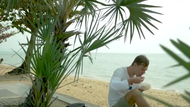 Мужчина ест тайский суп Том Ям сидит на пляже, 4k замедленной съемки — стоковое видео