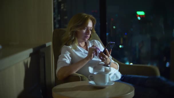 Mladá, krásná blonďatá žena pomocí smartphone, na židli v pokoji s panoramatickým výhledem na mrakodrapy v noci. 4k, rozostření pozadí. — Stock video