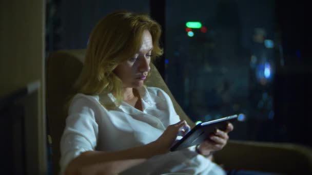 Молода красива блондинка використовує смартфон, на стільці в кімнаті з панорамним вікном з видом на хмарочоси вночі. 4k, розмити фон . — стокове відео