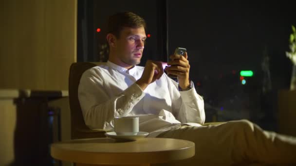 Mooie, jonge man, met behulp van een smartphone, op een stoel in een kamer met een panoramisch raam met uitzicht op de wolkenkrabbers in de nacht. 4k, wazig de achtergrond. — Stockvideo
