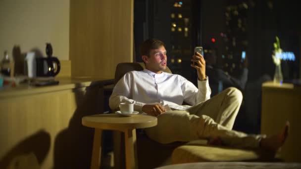 Joven y hermoso hombre usando un teléfono inteligente, en una silla en una habitación con una ventana panorámica con vistas a los rascacielos por la noche. 4k, borra el fondo . — Vídeos de Stock