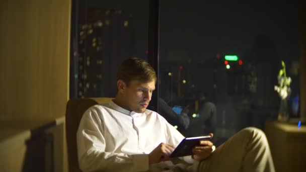 Joven y hermoso hombre usando un teléfono inteligente, en una silla en una habitación con una ventana panorámica con vistas a los rascacielos por la noche. 4k, borra el fondo . — Vídeo de stock