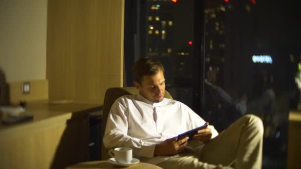 Joven y hermoso hombre usando un teléfono inteligente, en una silla en una habitación con una ventana panorámica con vistas a los rascacielos por la noche. 4k, borra el fondo . — Vídeo de stock