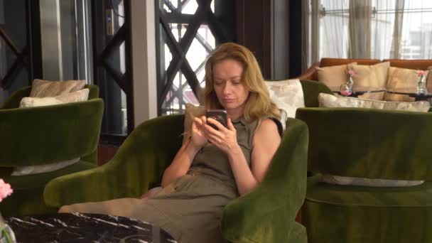Blond kvinna använder telefonen i ett café med utsikt från fönstret till skyskrapor. 4k — Stockvideo