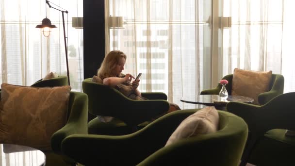 Blonďatá žena používá telefon v kavárně s výhledem z okna na mrakodrapy. 4k