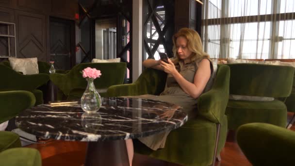Donna bionda usa il telefono in un caffè con vista dalla finestra ai grattacieli. 4k — Video Stock