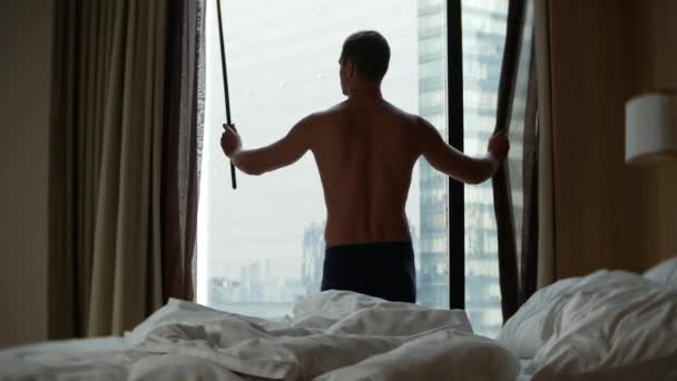 De man in korte broek sluit de gordijnen op het venster, het uitzicht uit het raam naar de wolkenkrabbers. 4k — Stockvideo