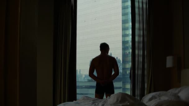 Un homme heureux en short se tient près de la fenêtre après le réveil, vue de la fenêtre aux gratte-ciel. 4k Mouvement lent — Video