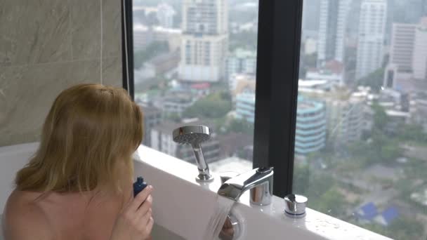 Красива жінка насолоджується розслаблюючою ванною в розкішній ванній кімнаті з вікном. Концепція способу життя та догляду за красою. вид з вікна на хмарочоси. 4k — стокове відео