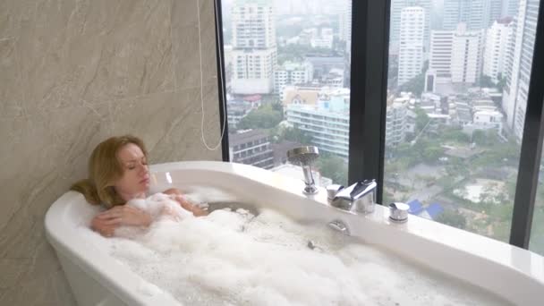 Krásná žena se těší relaxační koupel v luxusní koupelnu s oknem. Koncepce péče o životní styl a krása. pohled z okna na mrakodrapy. 4k