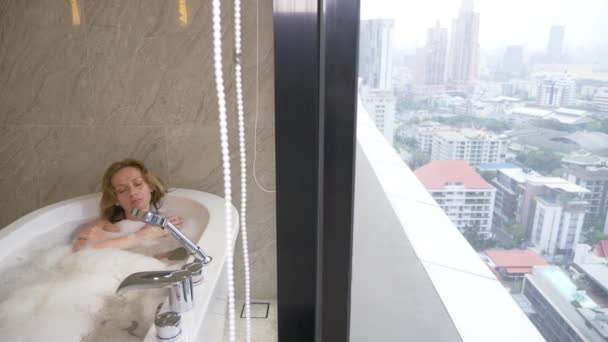 Hermosa mujer disfrutando de un baño relajante en un baño de lujo con ventana. Concepto de estilo de vida y cuidado de belleza. vista desde la ventana a los rascacielos. 4k — Vídeo de stock