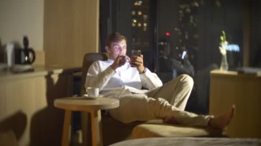 Genç, güzel bir sandalyeye gökdelenler geceleri bakan panoramik pencereli bir odada bir akıllı telefon kullanan adam. 4k, arka plan bulanıklık.
