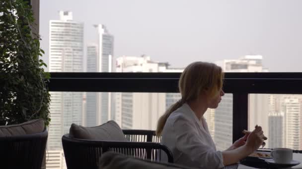 Hermosa chica elegante, desayunando en una cafetería en la terraza con una hermosa vista de los rascacielos. Una mujer tomando café caliente en la lujosa terraza de los hoteles. 4k , — Vídeo de stock