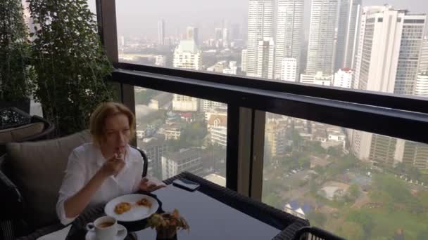 Szép elegáns lány, egy kávézó reggelizik a teraszon gyönyörű kilátás nyílik a felhőkarcolók. Egy nő forró kávét iszik a szállás luxus teraszon. 4k, — Stock videók