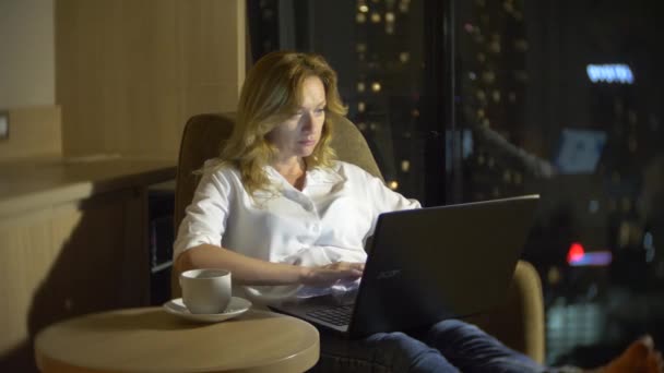 Junge, schöne blonde Frau mit Laptop, auf einem Stuhl in einem Raum mit einem Panoramafenster mit Blick auf die Wolkenkratzer in der Nacht. 4k, den Hintergrund verwischen — Stockvideo