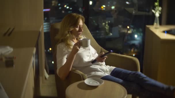 Joven, hermosa mujer rubia usando un panel de control de televisión, en una silla en una habitación con una ventana panorámica con vistas a los rascacielos por la noche. 4k, borra el fondo . — Vídeos de Stock