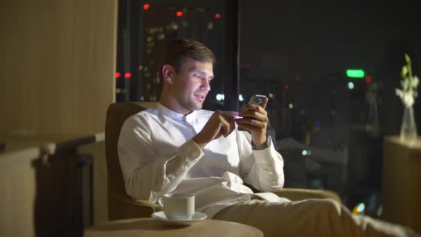 Joven y hermoso hombre usando un teléfono inteligente, en una silla en una habitación con una ventana panorámica con vistas a los rascacielos por la noche. 4k, borra el fondo . — Vídeo de stock