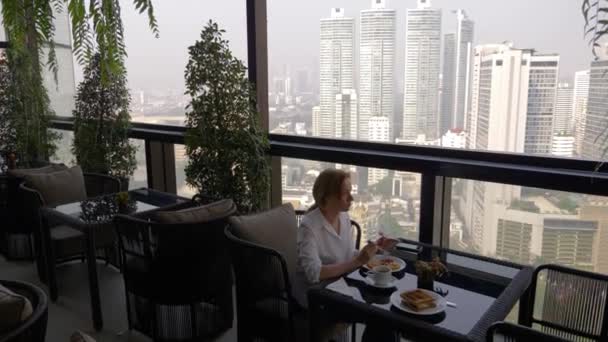 Bella ragazza elegante, facendo colazione in un caffè sulla terrazza con una splendida vista sui grattacieli. Una donna che beve caffè caldo sulla lussuosa terrazza dell'hotel. 4k , — Video Stock