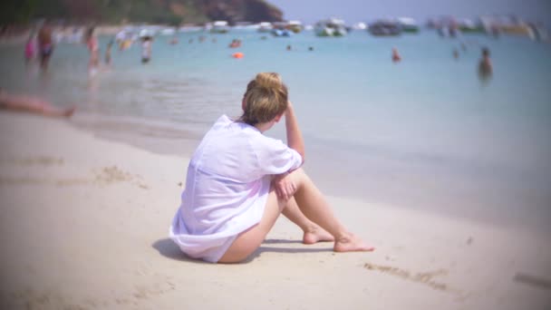 Une femme réfléchie et triste, assise seule sur le sable dans une plage bondée, 4k, au ralenti. flou de fond — Video