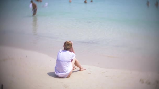 En eftertänksam, ledsen kvinna, sitter ensam på sanden i en fullpackad strand, 4k, slow motion. bakgrundsoskärpa — Stockvideo