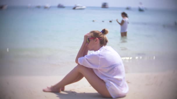 Задумчивая, грустная женщина, сидит одна на песке на переполненном пляже, 4К, замедленная съемка. Фоновое размытие — стоковое видео