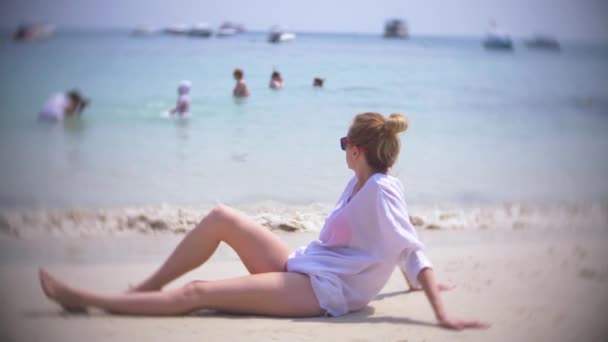 Une femme réfléchie et triste, assise seule sur le sable dans une plage bondée, 4k, au ralenti. flou de fond — Video