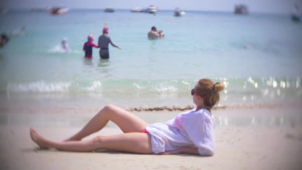 Une femme réfléchie et triste, assise seule sur le sable dans une plage bondée, 4k, au ralenti. flou de fond — Video