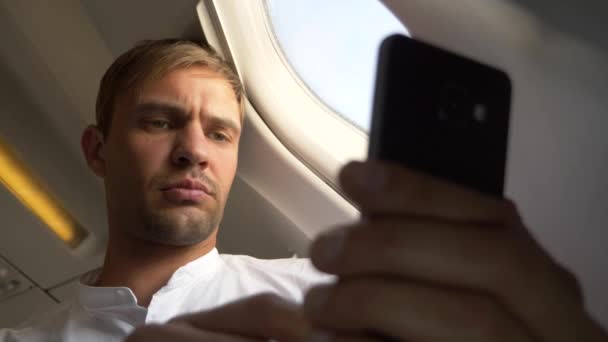 4k, γκρο πλαν. Πορτρέτο του ένας όμορφος νεαρός άνδρας που φαίνεται στο του smartphone, ενώ κάθονται δίπλα στο παράθυρο του αεροπλάνου κατά τη διάρκεια μιας πτήσης. — Αρχείο Βίντεο