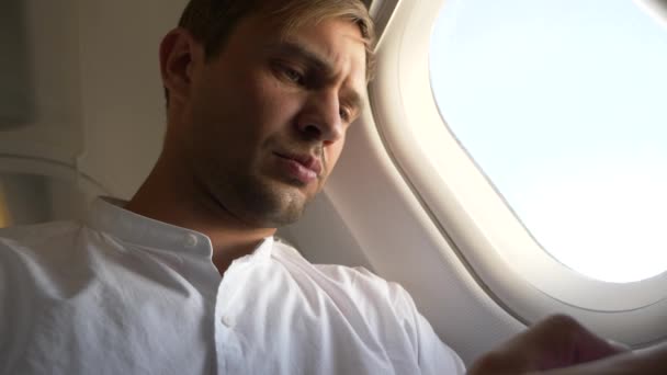 4k, close-up. Portrait of a handsome young man who looks at his smartphone while sitting by the window of an airplane during a flight. — Stock Video