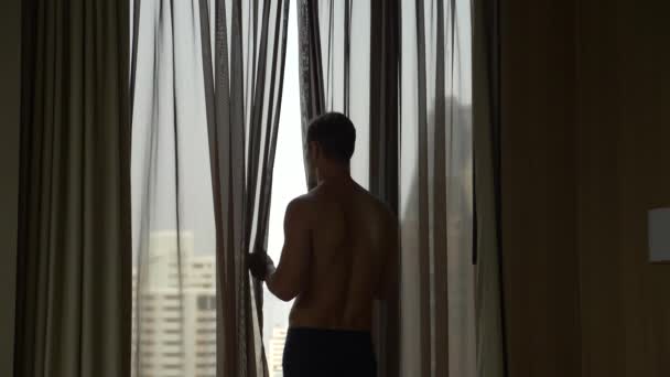 Ο άνθρωπος με το σορτς ανοίγει τις κουρτίνες στο παράθυρο, θέα από το παράθυρο τους ουρανοξύστες. 4 k αργή κίνηση — Αρχείο Βίντεο