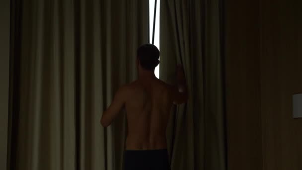 Homem de shorts abre as cortinas na janela, vista da janela para os arranha-céus. 4k câmera lenta — Vídeo de Stock