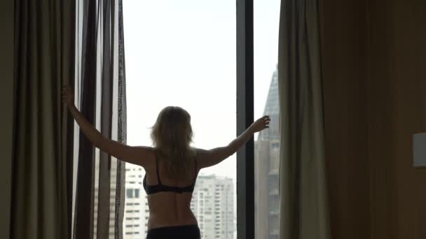 Ξανθιά γυναίκα στο εσωρούχων, κλείνει τις κουρτίνες στο παράθυρο, θέα από το παράθυρο τους ουρανοξύστες. 4 k αργή κίνηση — Αρχείο Βίντεο