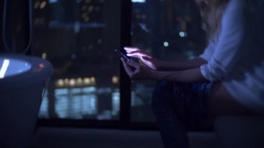 Bir telefon ve çalışma ile gece lüks tuvalet panoramik pencereli tuvalette oturan bir kadın. 4k