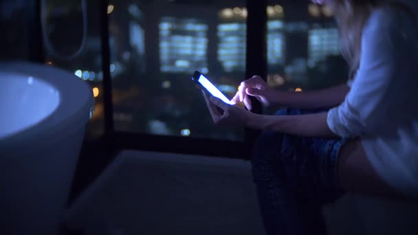 Uma mulher sentada no banheiro em um banheiro luxuoso com uma janela panorâmica à noite com um telefone e trabalhando. 4k — Vídeo de Stock