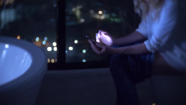 Женщина сидит на унитазе в роскошном туалете с панорамным окном ночью с телефоном и работает. 4k — стоковое видео
