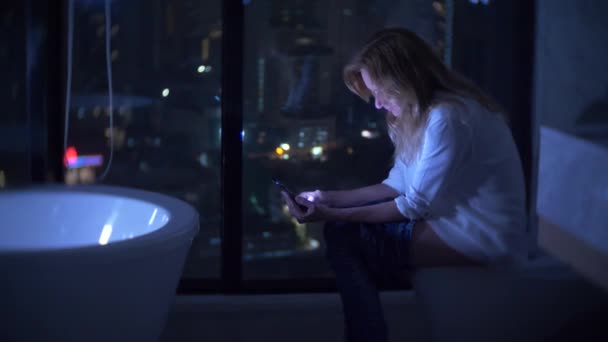 Жінка сидить на туалеті в розкішній туалеті з панорамним вікном вночі з телефоном і роботою. 4k — стокове відео