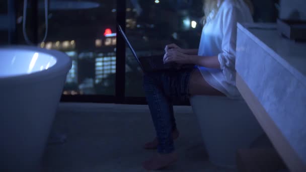 Uma mulher está sentada no banheiro em um banheiro luxuoso com uma janela panorâmica à noite com um laptop e funciona. 4k — Vídeo de Stock