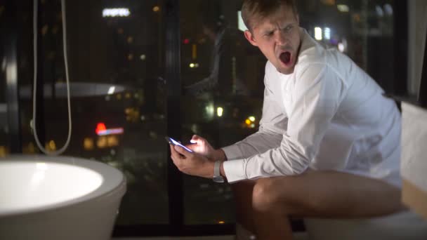 Un homme assis sur les toilettes dans des toilettes luxueuses avec une fenêtre panoramique la nuit avec un téléphone et de travail. 4k — Video