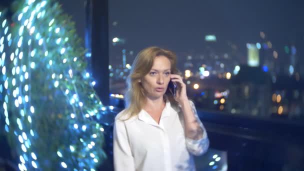 Jeune, belle femme blonde utilisant un smartphone, sur une terrasse du bar surplombant les gratte-ciel la nuit. 4k, flou de fond . — Video