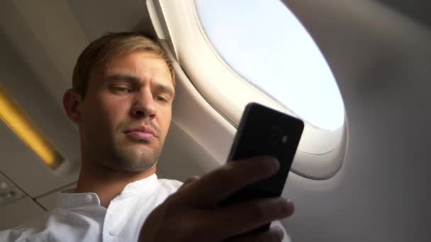 4k, primer plano. Retrato de un joven guapo que mira su teléfono inteligente mientras está sentado junto a la ventana de un avión durante un vuelo . — Vídeo de stock