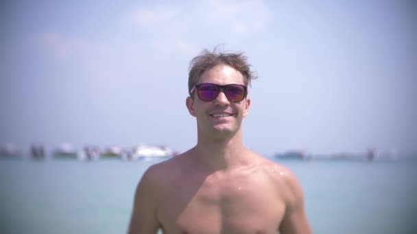 4k, slow motion, молодой человек стоит в море в солнцезащитных очках, брызгает водой и улыбается — стоковое видео