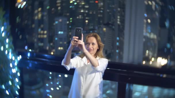 Jonge, mooie blonde vrouw met behulp van een smartphone, op een bar terras met uitzicht op de wolkenkrabbers in de nacht. 4k, achtergrond vervagen. — Stockvideo