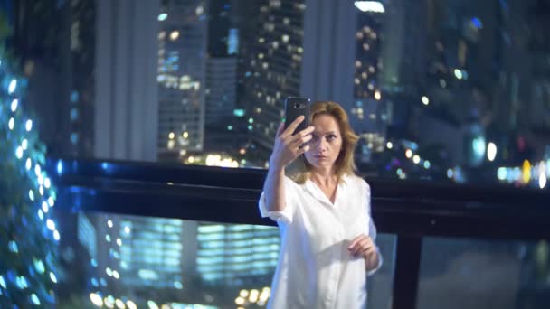 Jeune, belle femme blonde utilisant un smartphone, sur une terrasse du bar surplombant les gratte-ciel la nuit. 4k, flou de fond . — Video