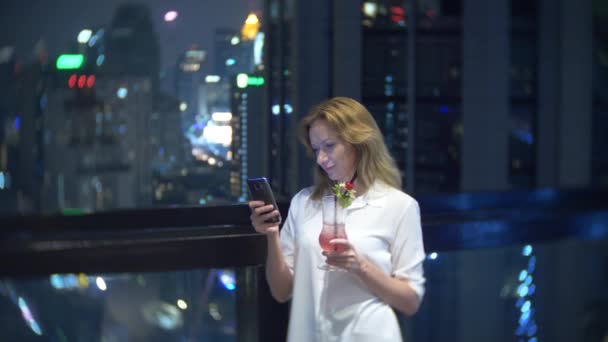 Joven rubia relajándose y bebiendo cóctel en el bar con vistas a los rascacielos por la noche. 4k, desenfoque de fondo — Vídeos de Stock