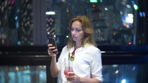 Joven rubia relajándose y bebiendo cóctel en el bar con vistas a los rascacielos por la noche. 4k, desenfoque de fondo — Vídeos de Stock