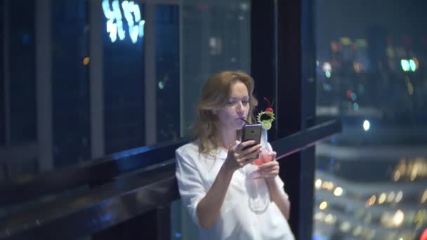 Jeune femme blonde relaxante et buvant un cocktail au bar avec vue sur les gratte-ciel la nuit. 4k, flou de fond — Video