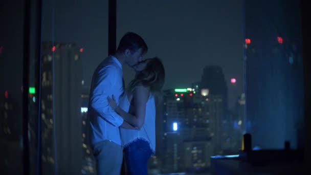 Молодая пара целуется на фоне панорамного окна с видом на город. Вечернее время. 4K, размытие фона — стоковое видео