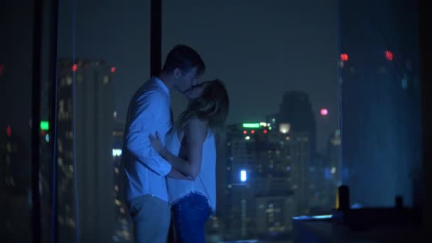 Um jovem casal está se beijando no fundo de uma janela panorâmica com vista para a cidade. Boa noite, à noite. 4k, borrão de fundo — Vídeo de Stock