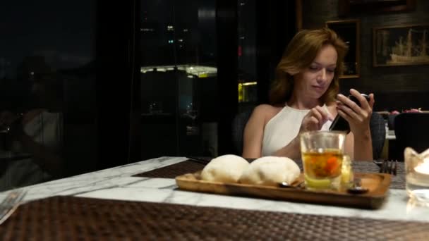 Wanita cantik dengan telepon di malam hari menunggu di restoran. 4k, kabur latar belakang — Stok Video